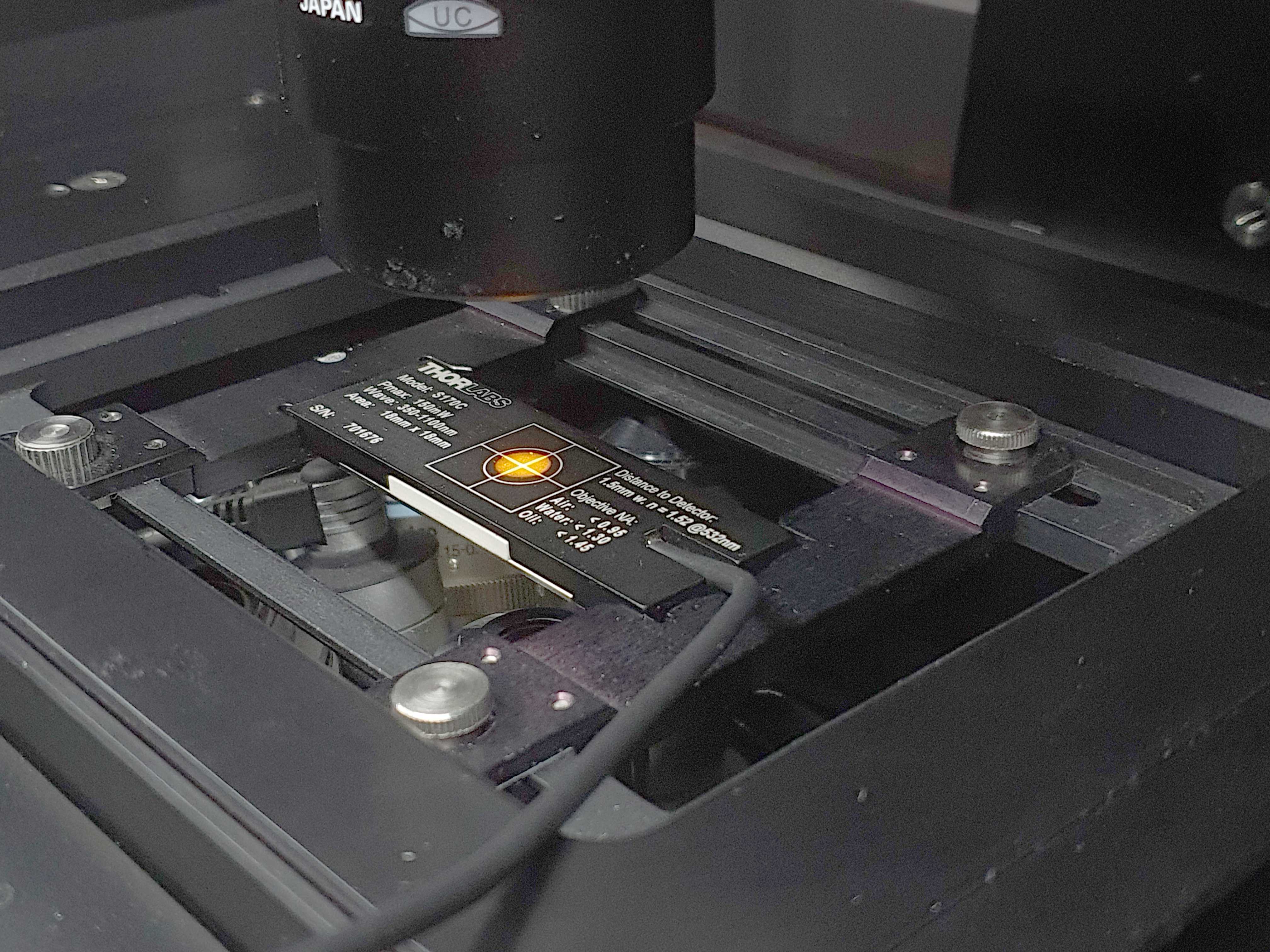 Figure 4. Slide power meter sensor mounted on the stage of an inverted microscope and aligned to the center of the sensing area using transmitted light and the cross mark located at the back of the sensor. Figure 4. Slide power meter sensor mounted on the stage of an inverted microscope and aligned to the center of the sensing area using transmitted light and the cross mark located at the back of the sensor.